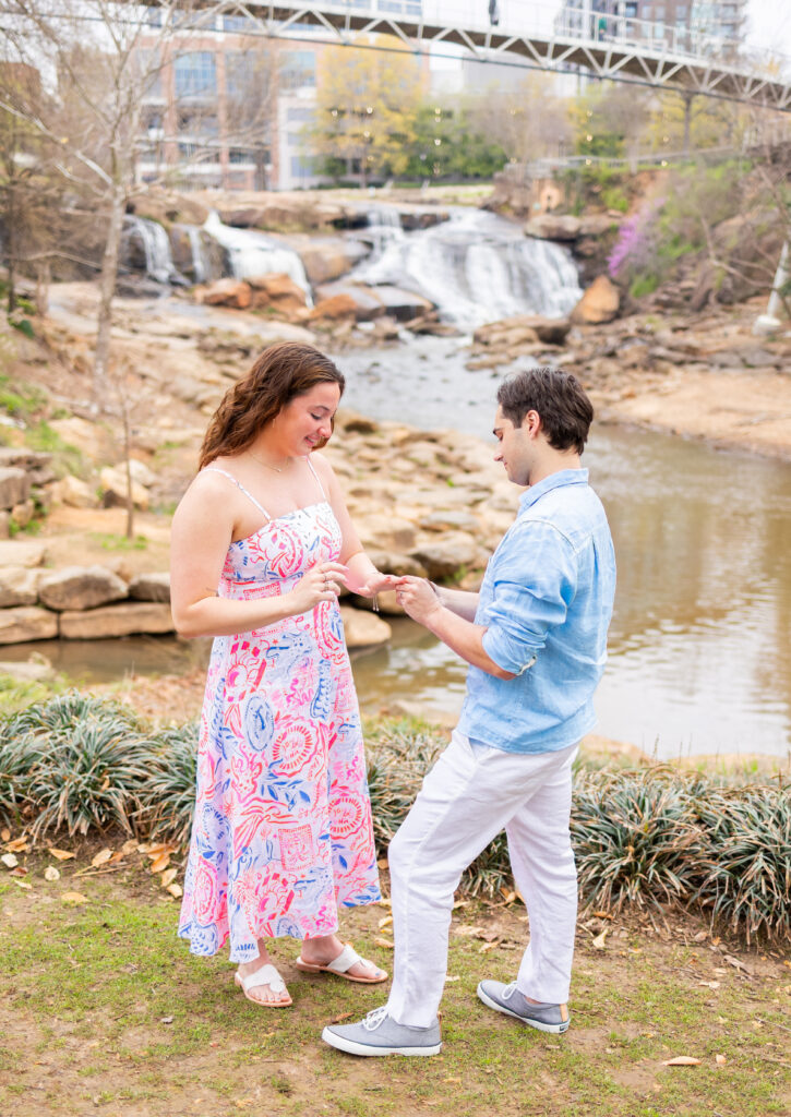 guy putting ring on girls finger after proposal in front of river in greenville